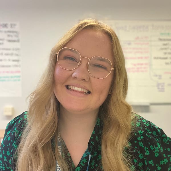 A blond woman wearing glasses smiles in front of a whiteboard.