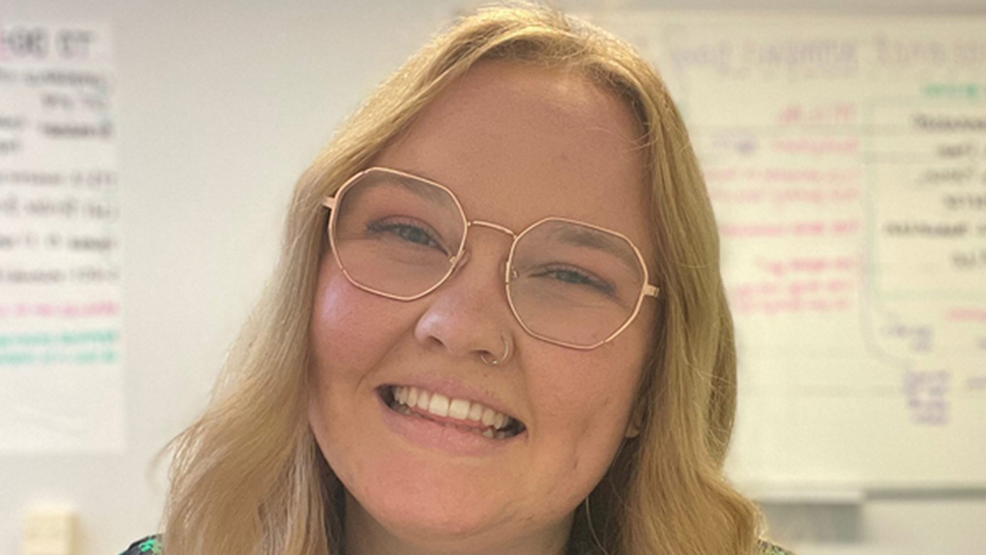 A blond woman wearing glasses smiles in front of a whiteboard.